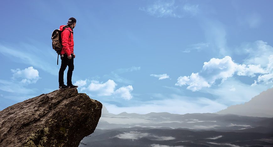 Mann mit roter Jacke und Rucksack steht auf Bergipfel und schaut in die Ferne